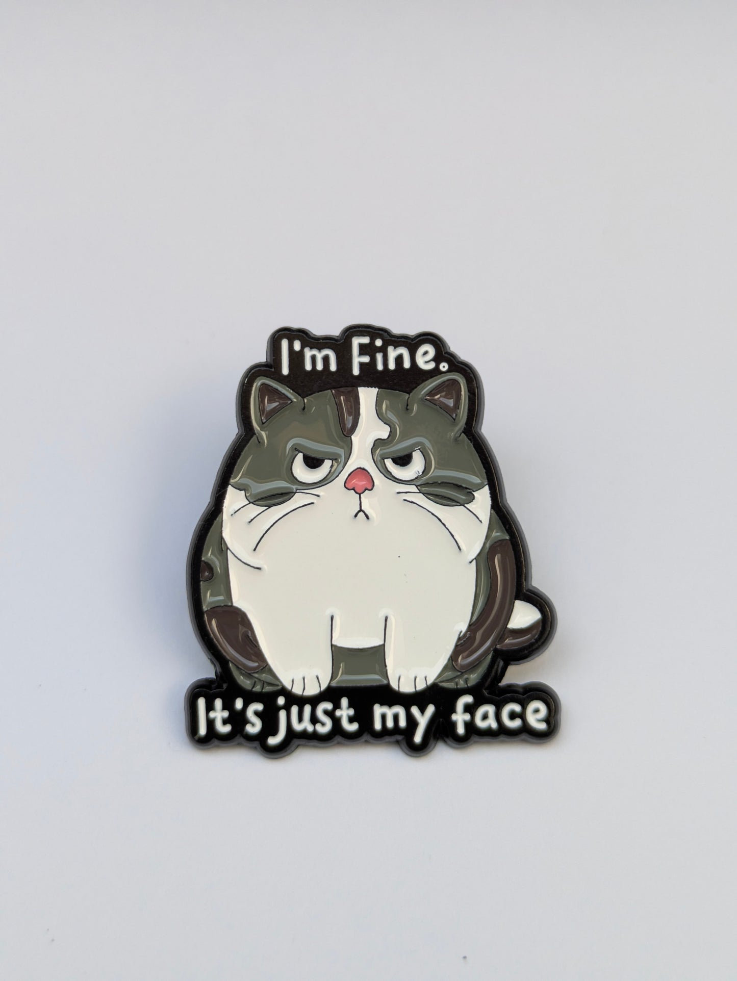 Cat-shaped enamel pin with text 'I'm Fine, It's just my face' on a white background