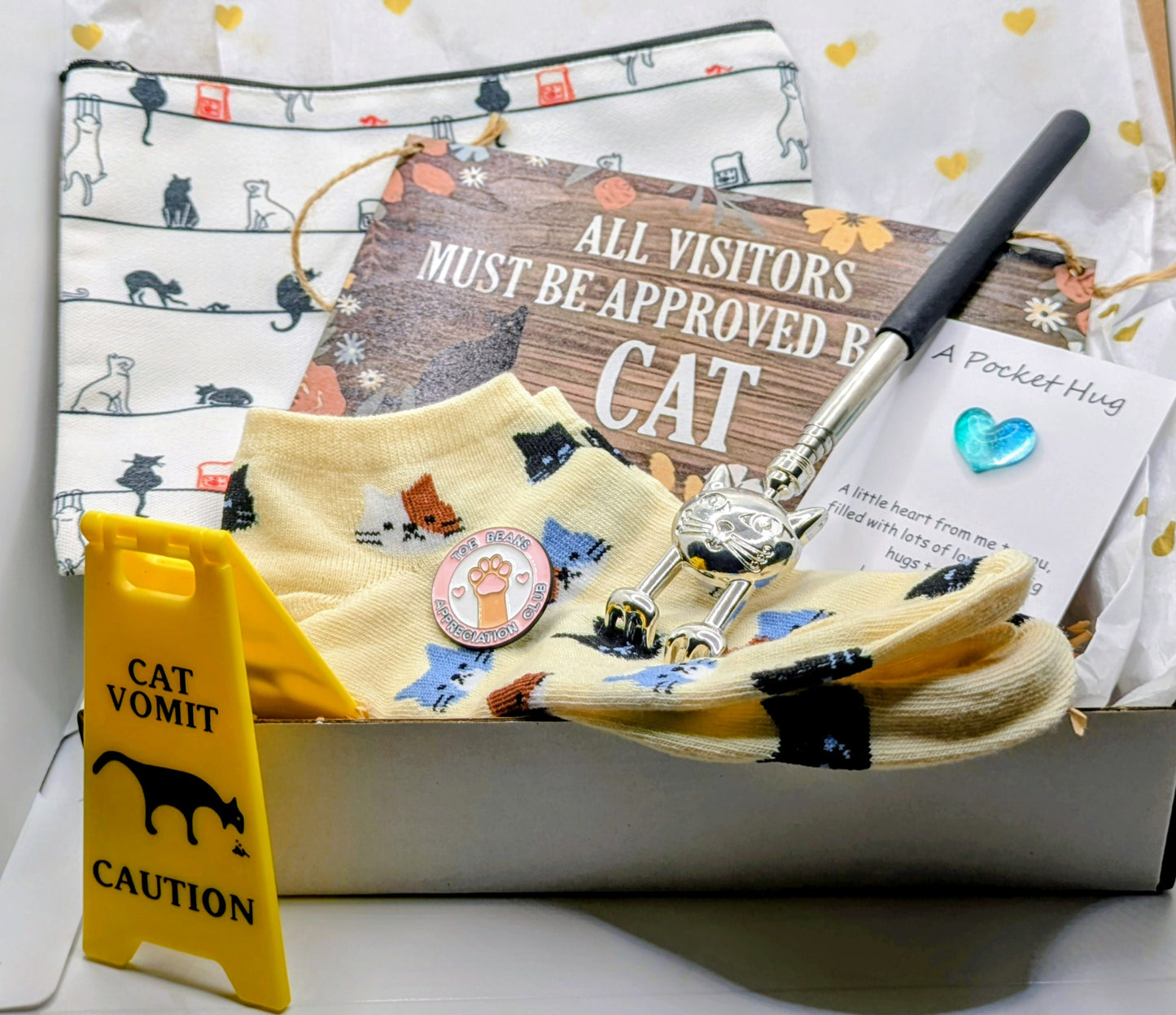 A collection of cat-themed gifts in a box. Features a yellow warning sign saying "Cat Vomit", socks with cat designs, a sign reading "All Visitors Must Be Approved By Cat", a cat-shaped back scratcher, a pocket hug, and a make-up bag with cat illustrations.