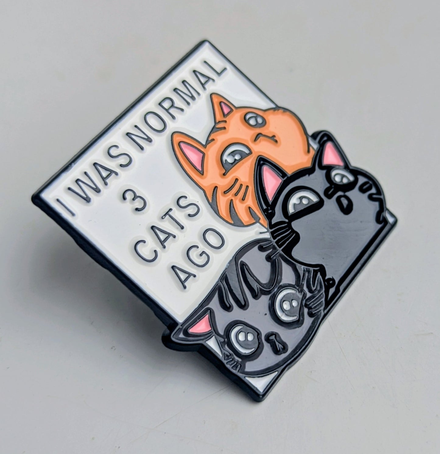 Enamel pin with cartoon cats and text 'I was normal 3 cats ago' on a gray background