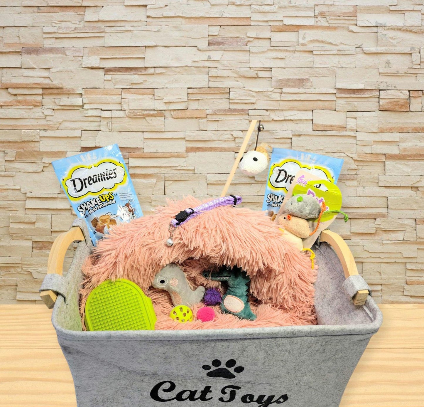 Grey fabric cat toy storage box filled with pink plush bed, assorted cat toys, and treat packets.