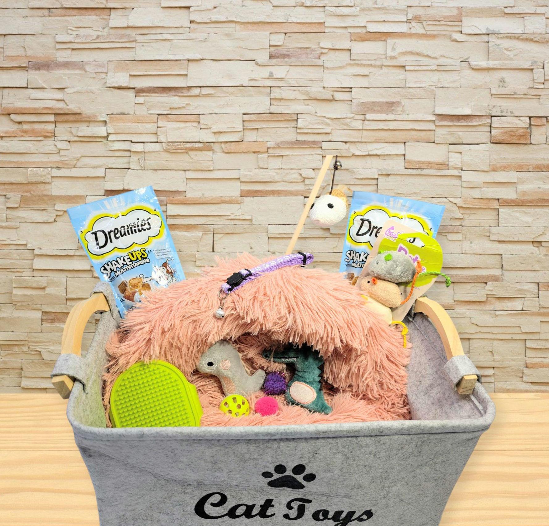 Grey fabric cat toy storage box filled with pink plush bed, assorted cat toys, and treat packets.