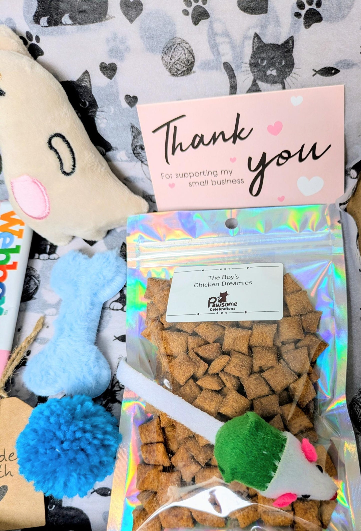 Mini cat treat bag with blue pom-pom toy, fabric mouse toy, and thank you card on paw-print fabric background.