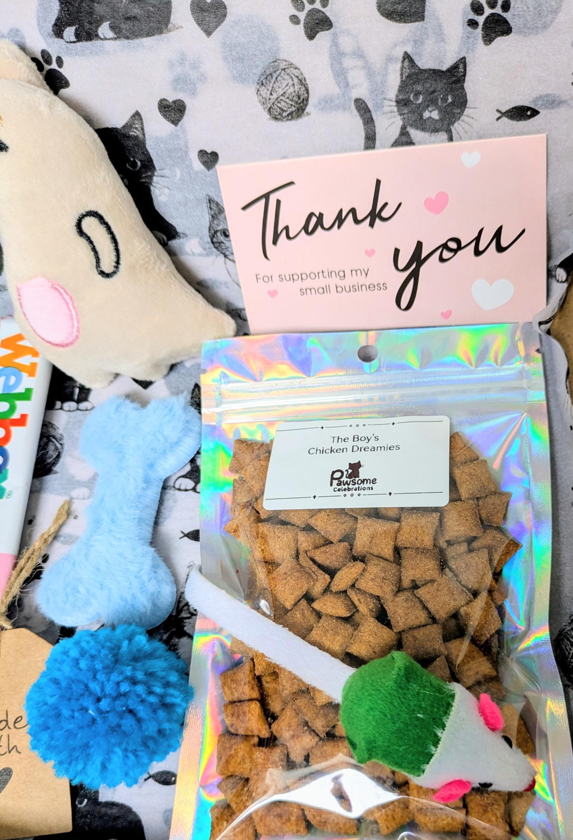 Mini cat treat bag with blue pom-pom toy, fabric mouse toy, and thank you card on paw-print fabric background.
