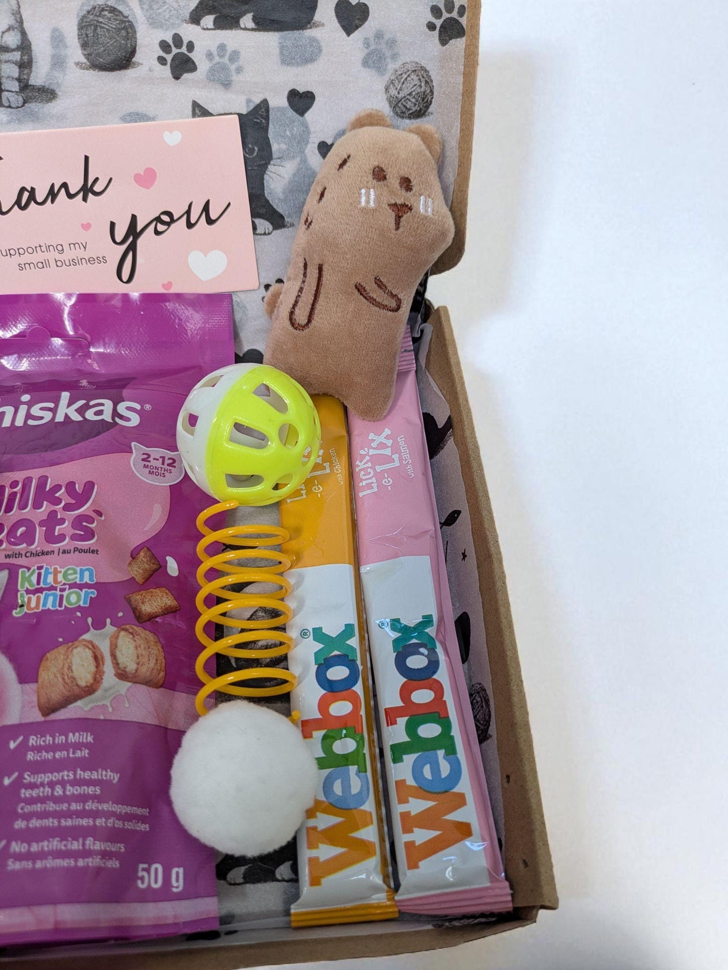 Open cardboard box with pink packet of Whiskas Milky Treats, a yellow and green ball toy, a brown mouse toy, yellow spring.