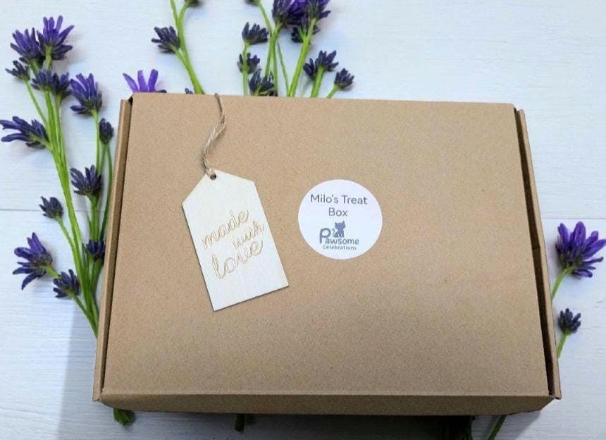 Brown cardboard cat treat box with white tag and round sticker, surrounded by purple flowers.