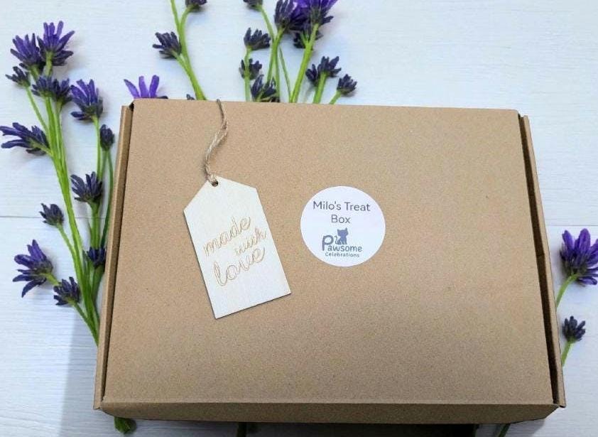 Brown cardboard gift box with white round label and hanging tag reading "Handmade with love" surrounded by purple flowers.