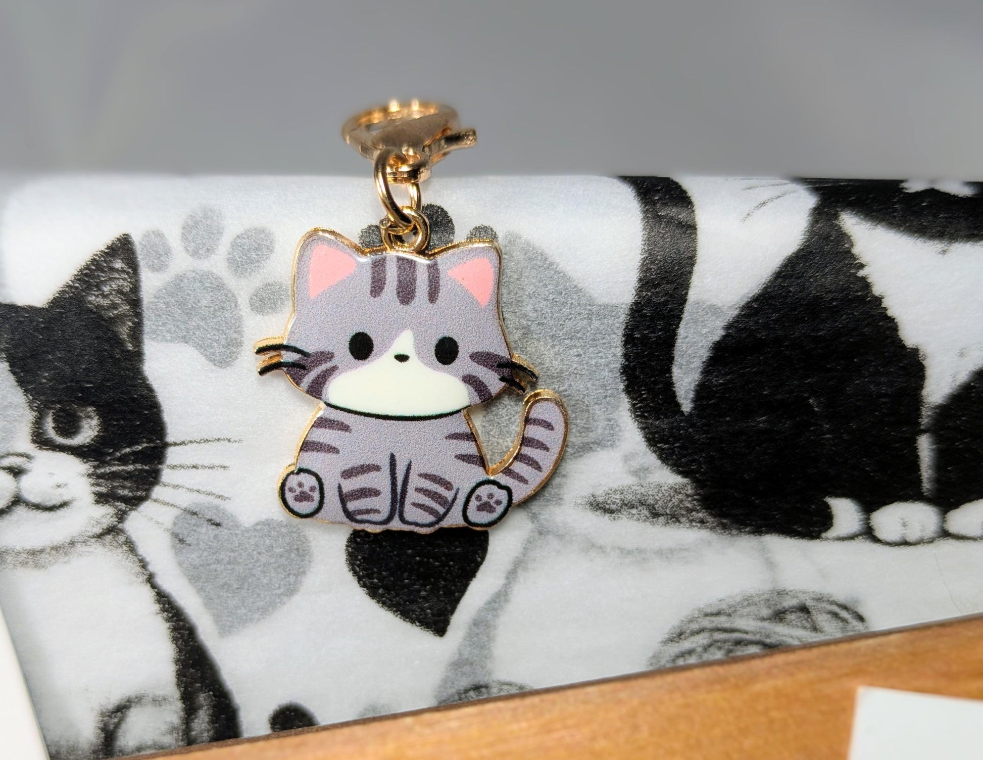 Cat-themed enamel keychain with a pink and gray tabby cat design on a black and white cat print fabric background.