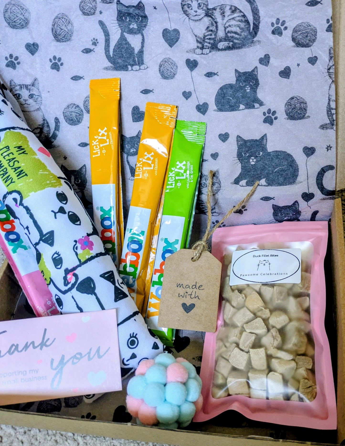 Pink gift box with cat treats, colorful catnip kicker ball, three cat treat sticks, and a thank you card on a cat-patterned.