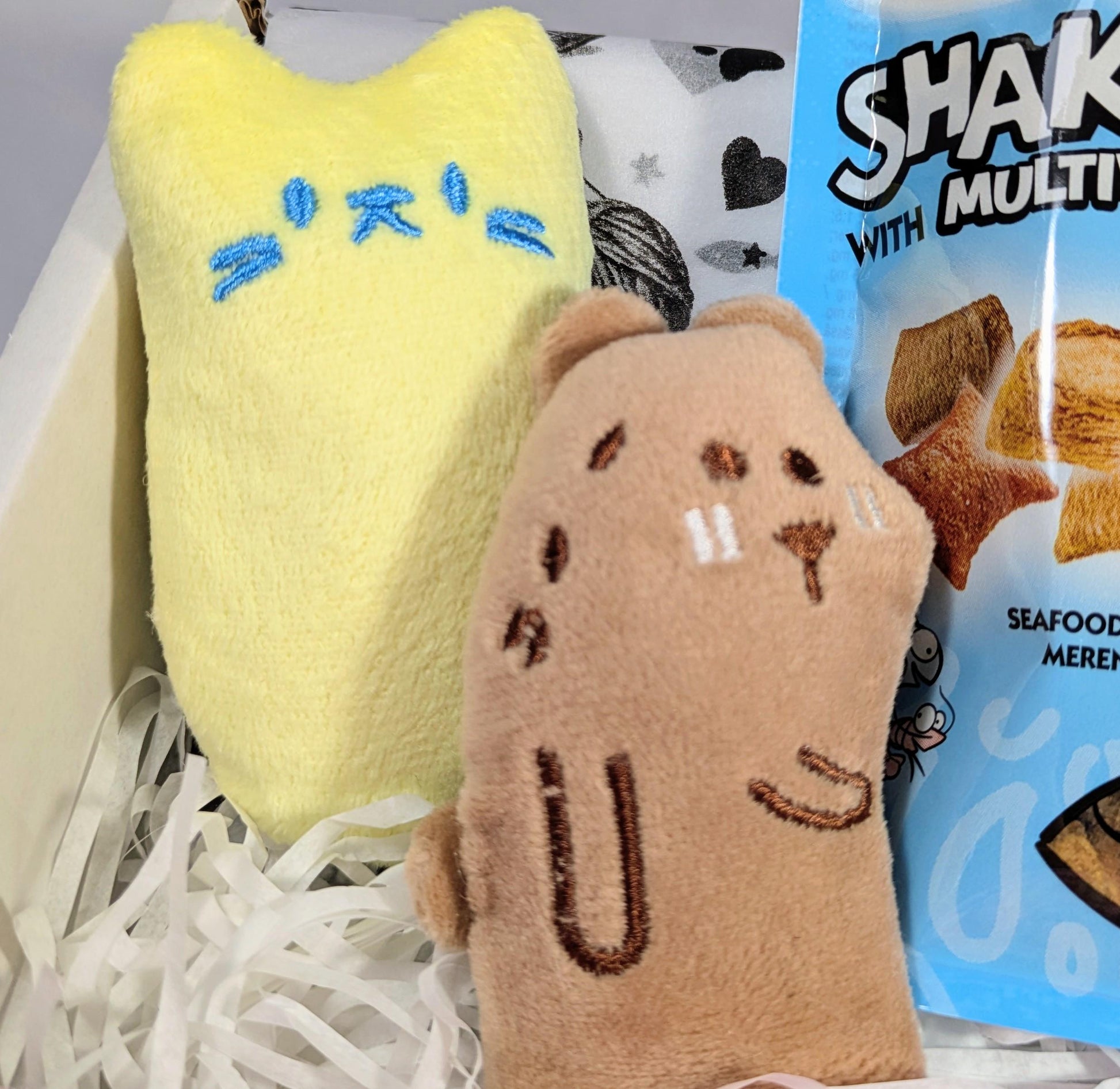 Yellow cat-shaped soft blanket with blue embroidery and brown plush toy with embroidered face inside white shredded paper.