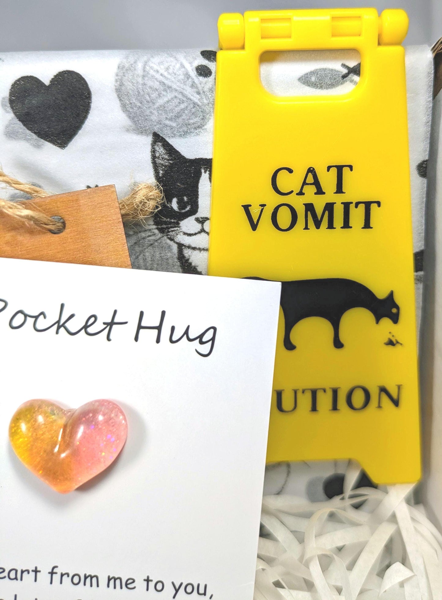 Yellow "Cat Vomit" caution sign, black cat silhouette, heart-shaped glass keepsake, and cat-themed fabric background.