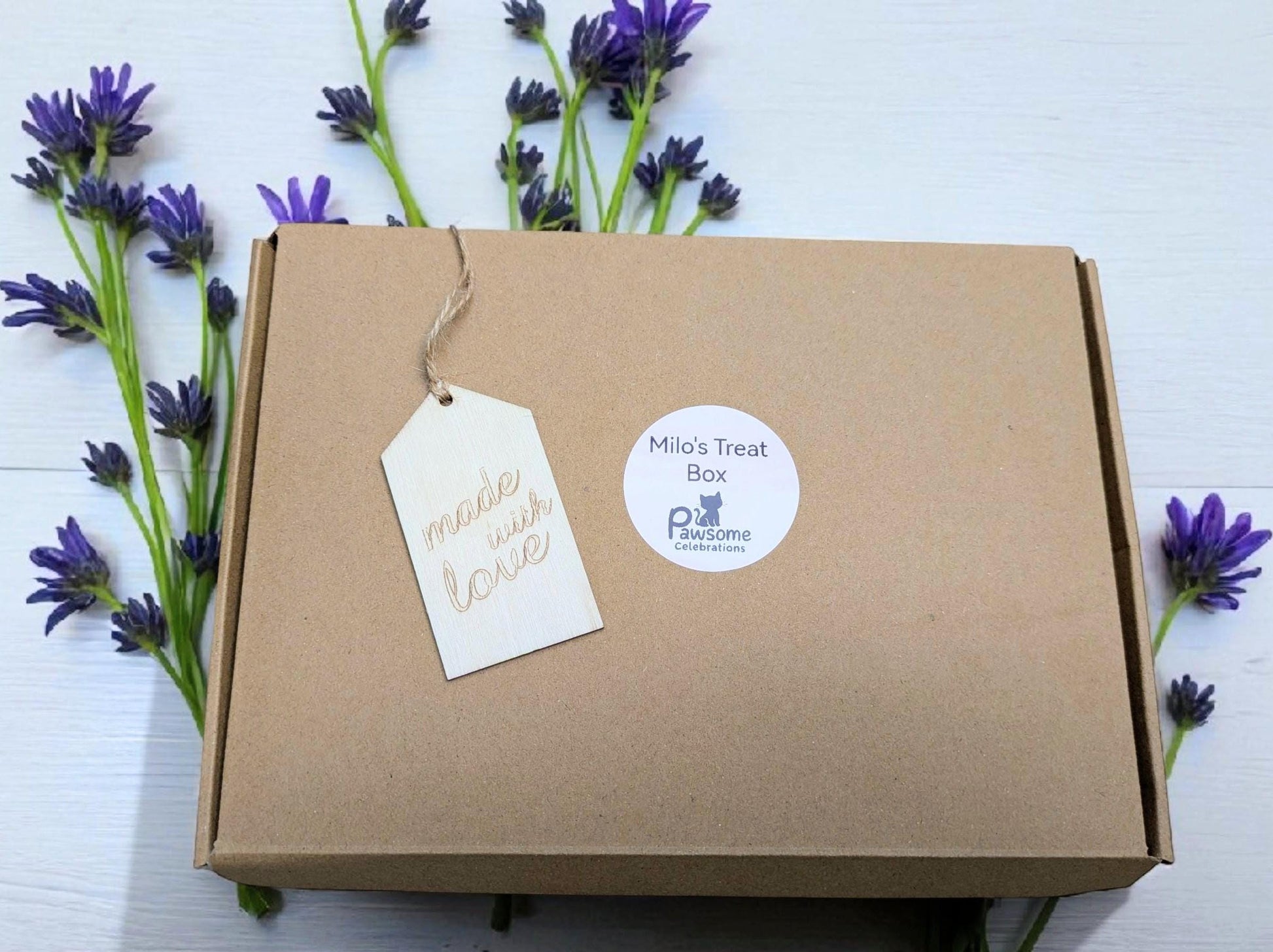 Brown cardboard gift box with white tags labeled "Natural Love" and "Mia's Treat," surrounded by purple flowers.