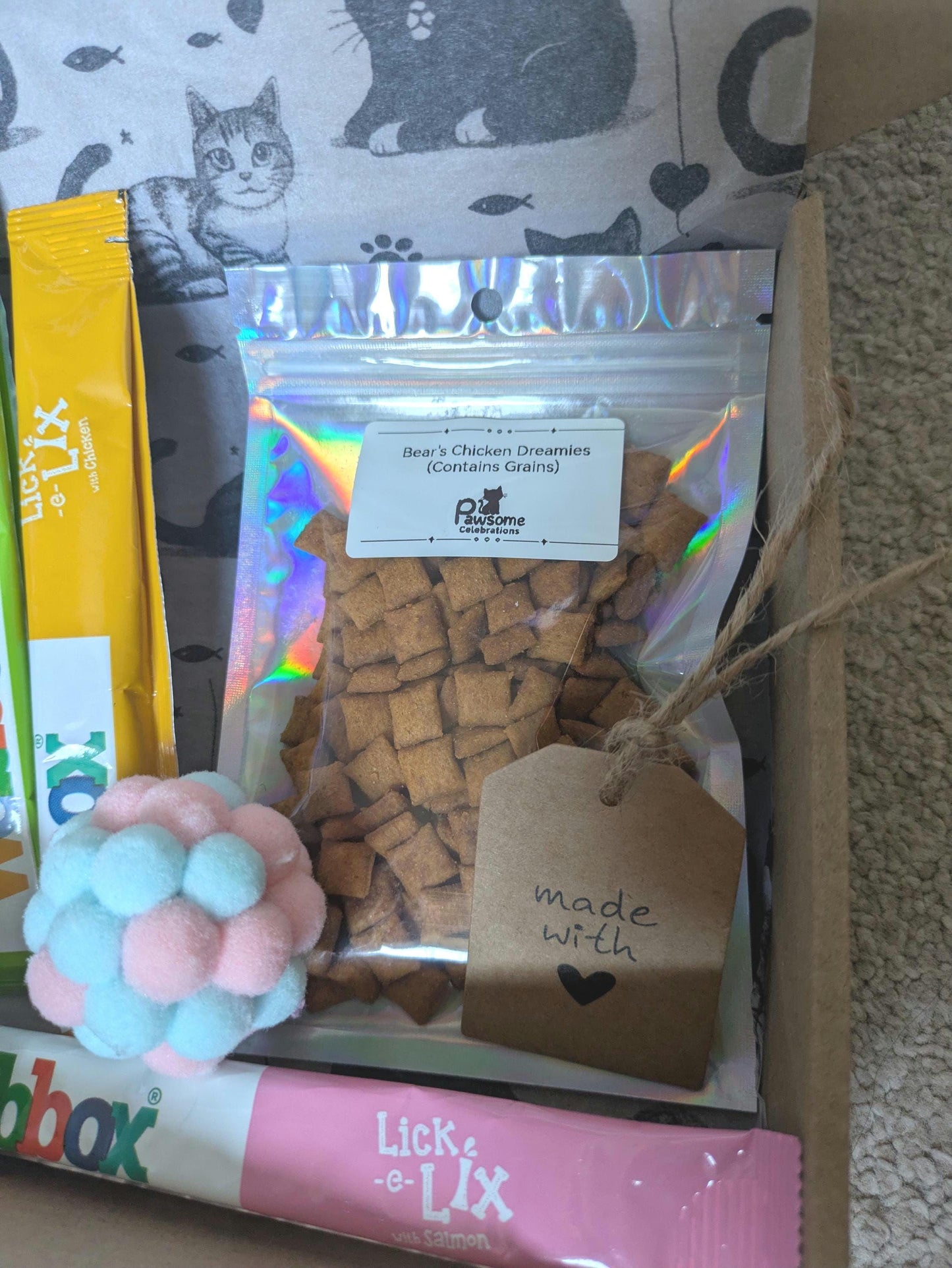 Small clear bag of cubed cat treats with personalized tag, pom-pom cat toy, and pink Lick a Lix stick in gift box.