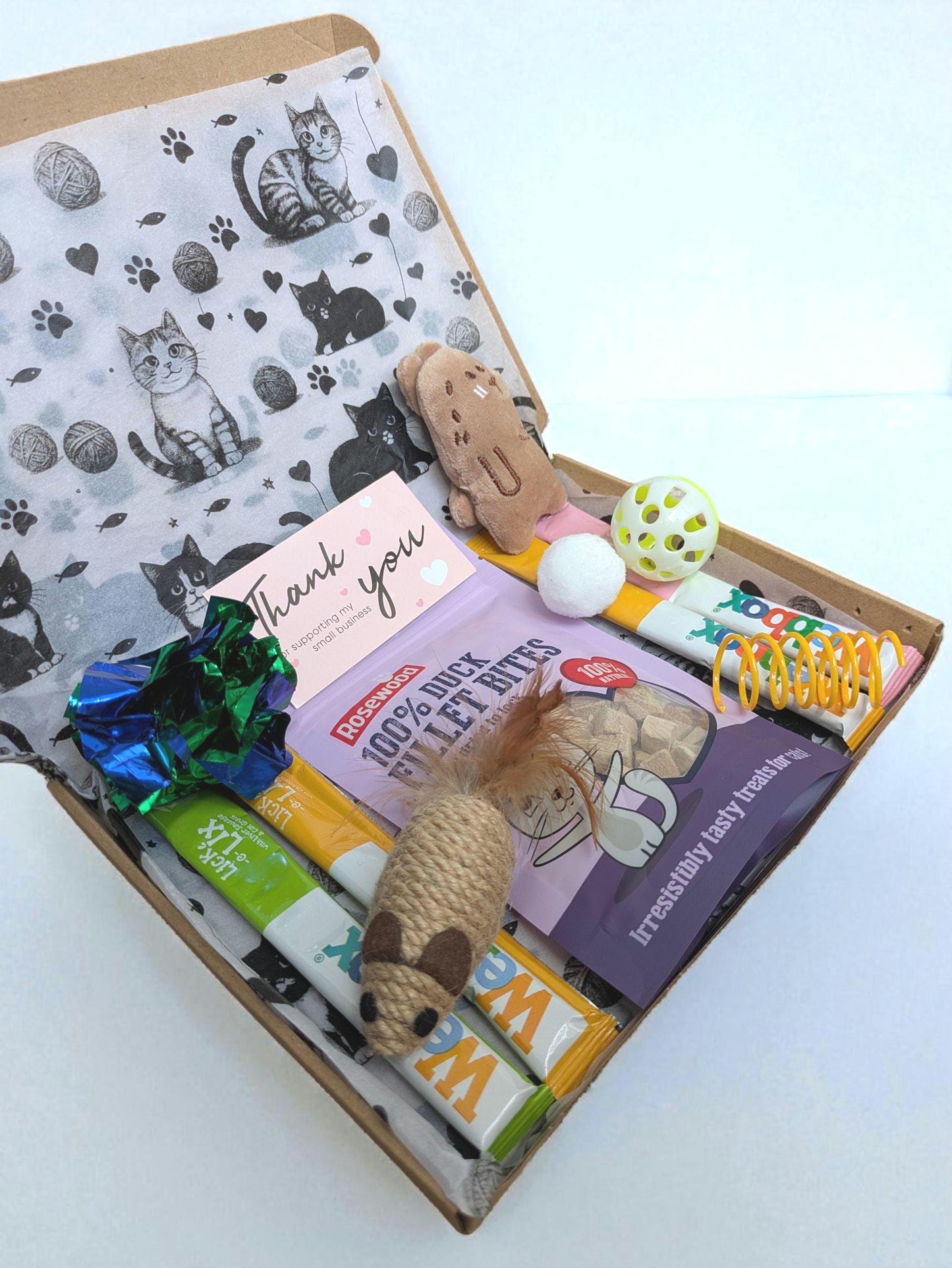 Open brown cardboard box with cat-themed tissue paper, catnip toys, and grain-free cat treat packets inside.