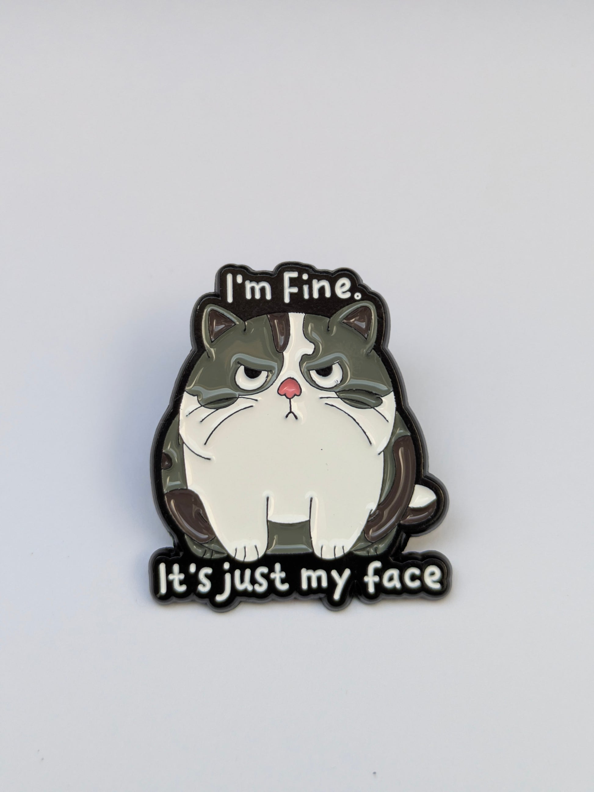 Cat-shaped enamel pin with text 'I'm Fine, It's just my face' on a white background