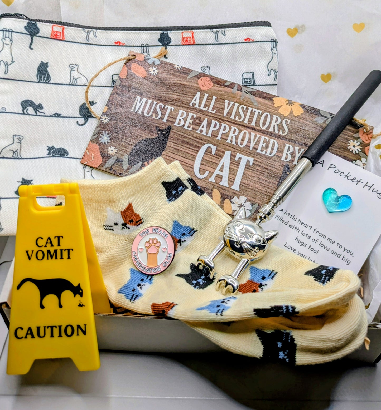 A collection of cat-themed gifts in a box. Features a yellow warning sign saying "Cat Vomit", socks with cat designs, a sign reading "All Visitors Must Be Approved By Cat", a cat-shaped back scratcher, and a make-up bag with cat illustrations.