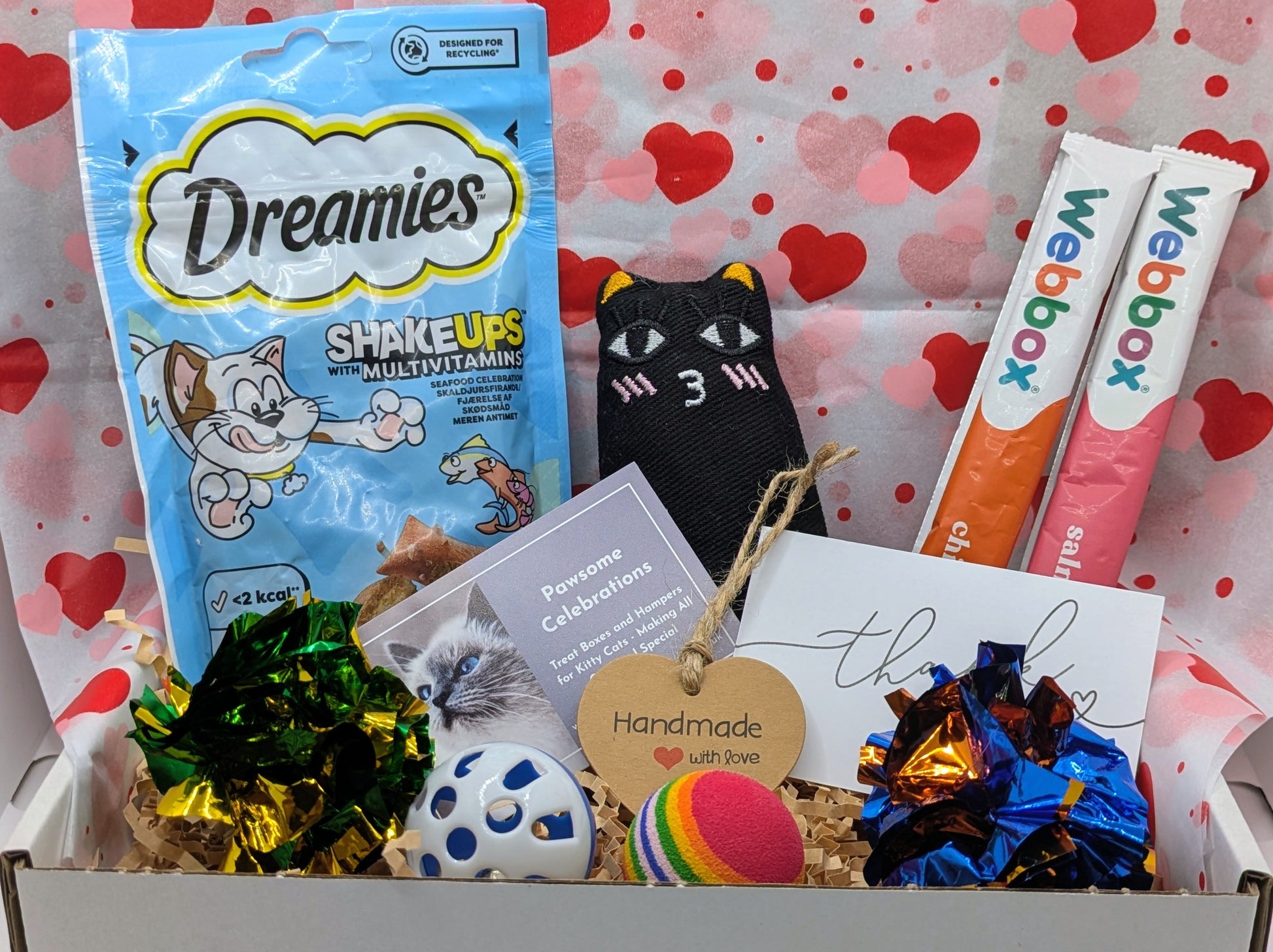 Cat-themed gift box with Dreamies cat treats, Weebly products, and decorative items on a heart-patterned background.
