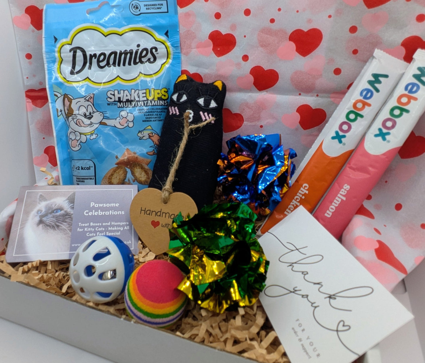 Assorted pet products including a bag of Dreamies cat treats, a toy, and a card on a heart-patterned background.
