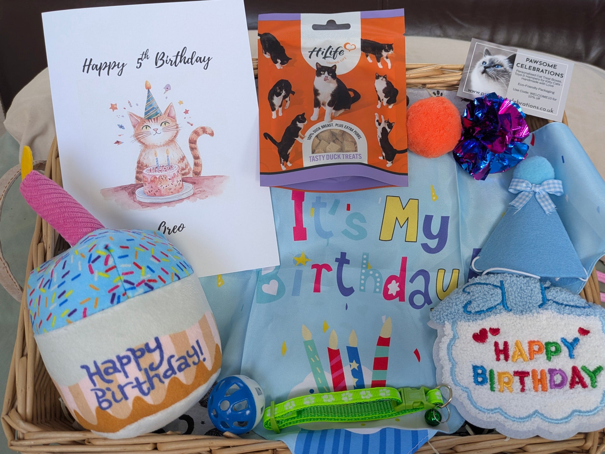 Cat birthday gift basket with toys, card, and treats on a table.