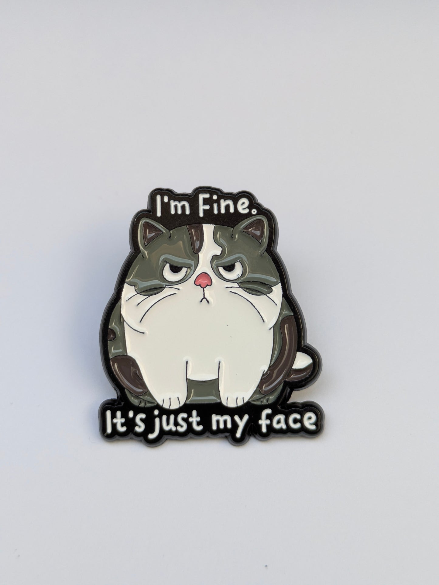 Cat-shaped enamel pin with text 'I'm Fine. It's just my face' on a white background