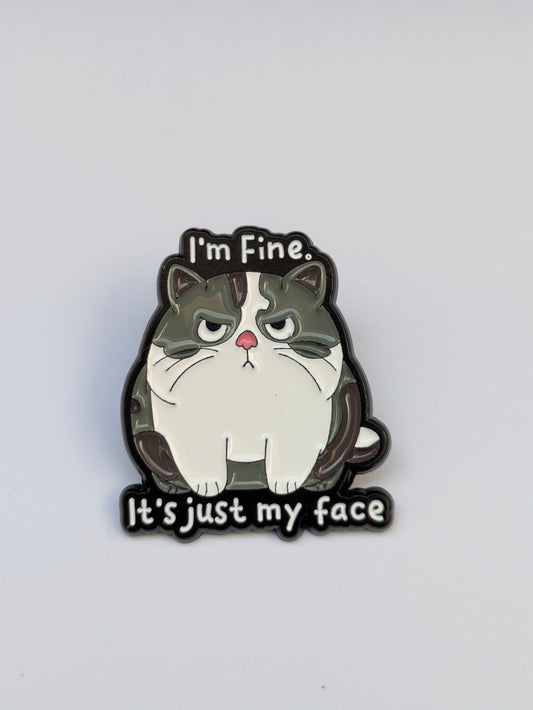 Cat-shaped enamel pin with text 'I'm Fine. It's just my face' on a white background