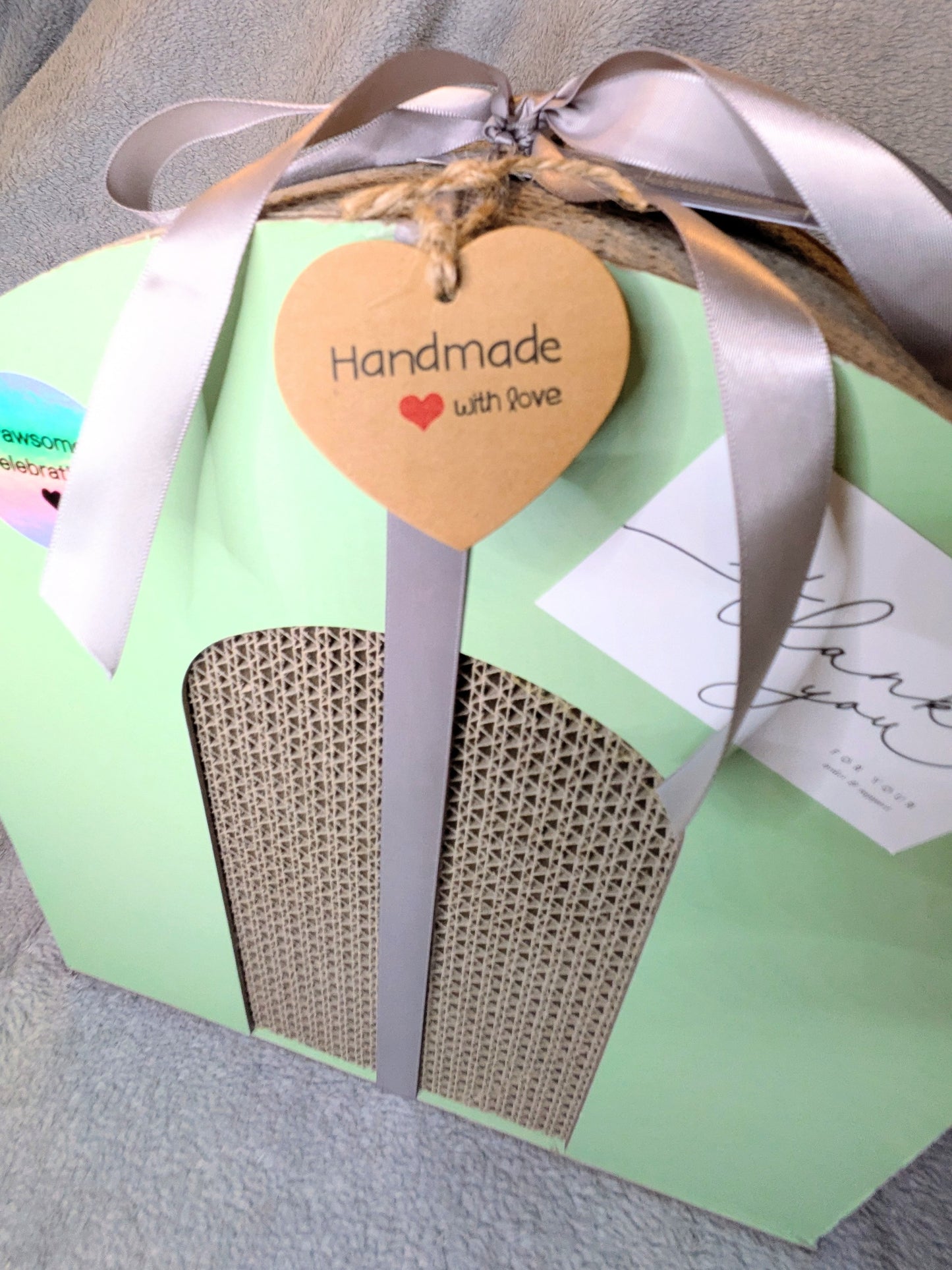 Foldable cat scratcher with a heart-shaped tag reading 'Handmade with Love' on a grey surface.