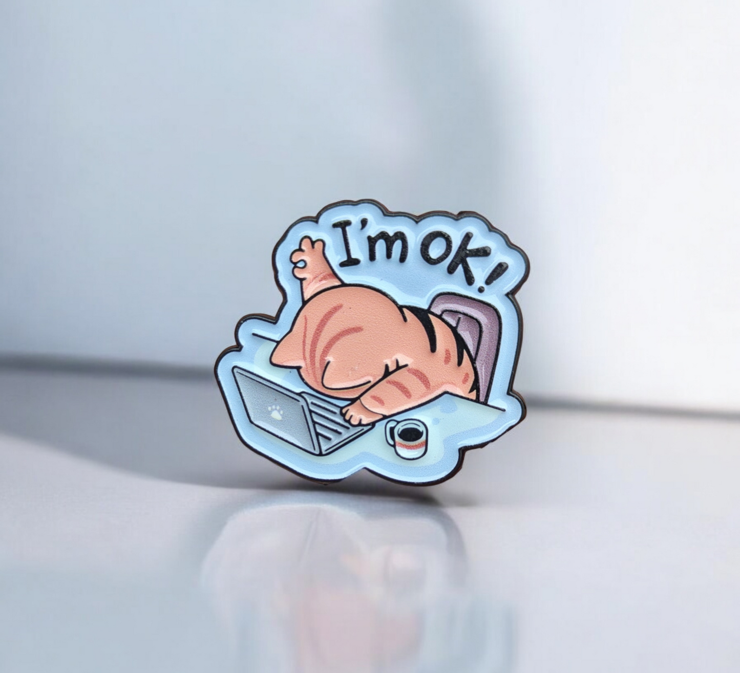 Enamel pin with a cartoon cat in a bubble saying 'I'm ok!' on a light blue background