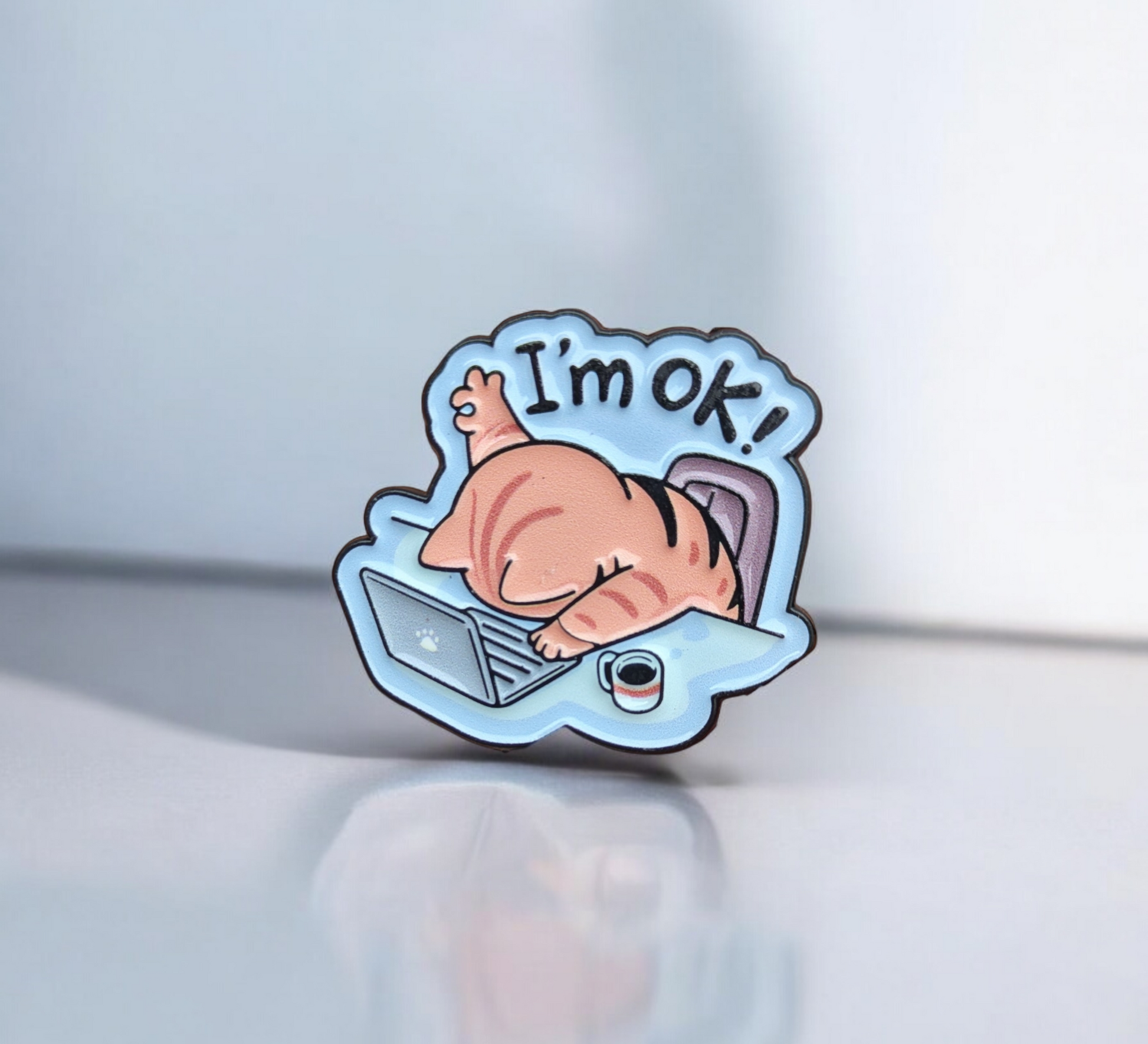 Enamel pin with a cartoon cat in a bubble saying 'I'm ok!' on a light blue background