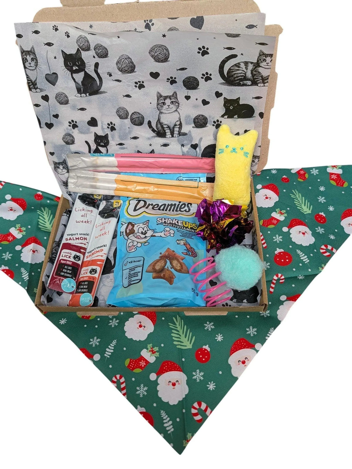 A Christmas-themed pet gift box filled with treats, toys, and accessories, set on a festive green bandana with Santa and snowflake patterns. The box is lined with white paper featuring black cat illustrations, and the contents include a blue bag of Dreamies dog treats, a yellow plush toy, a pink cat toy, and other holiday treats.