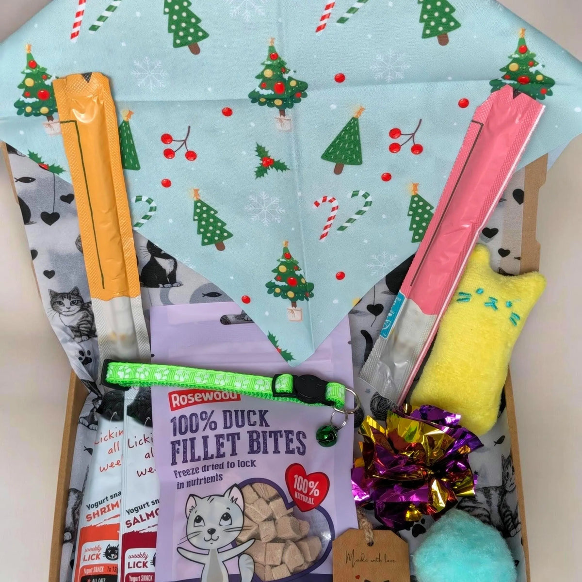 A curated Christmas-themed pet gift box filled with various treats and accessories. Visible items include a light blue cat-themed blanket with a Christmas tree pattern, a bag of Rosewoodu 100% Duck Fillet Bites, a yellow plush toy, a pink and white striped treat bag, a green collar, and a purple decorative bow.