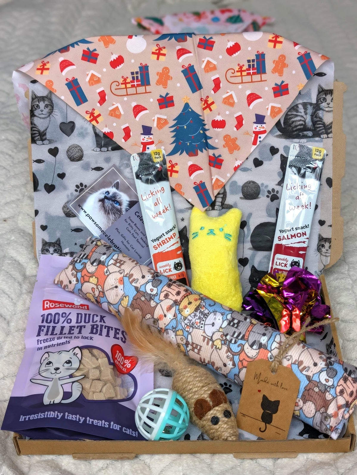 A curated pet gift box filled with treats, toys, and accessories, including a bag of 100% Duck Fillet Bites for cats, a yellow plush toy, a patterned pet toy roll, a blue ball, and a personalized tag, arranged in a cardboard box with a festive printed cat neck scarf.