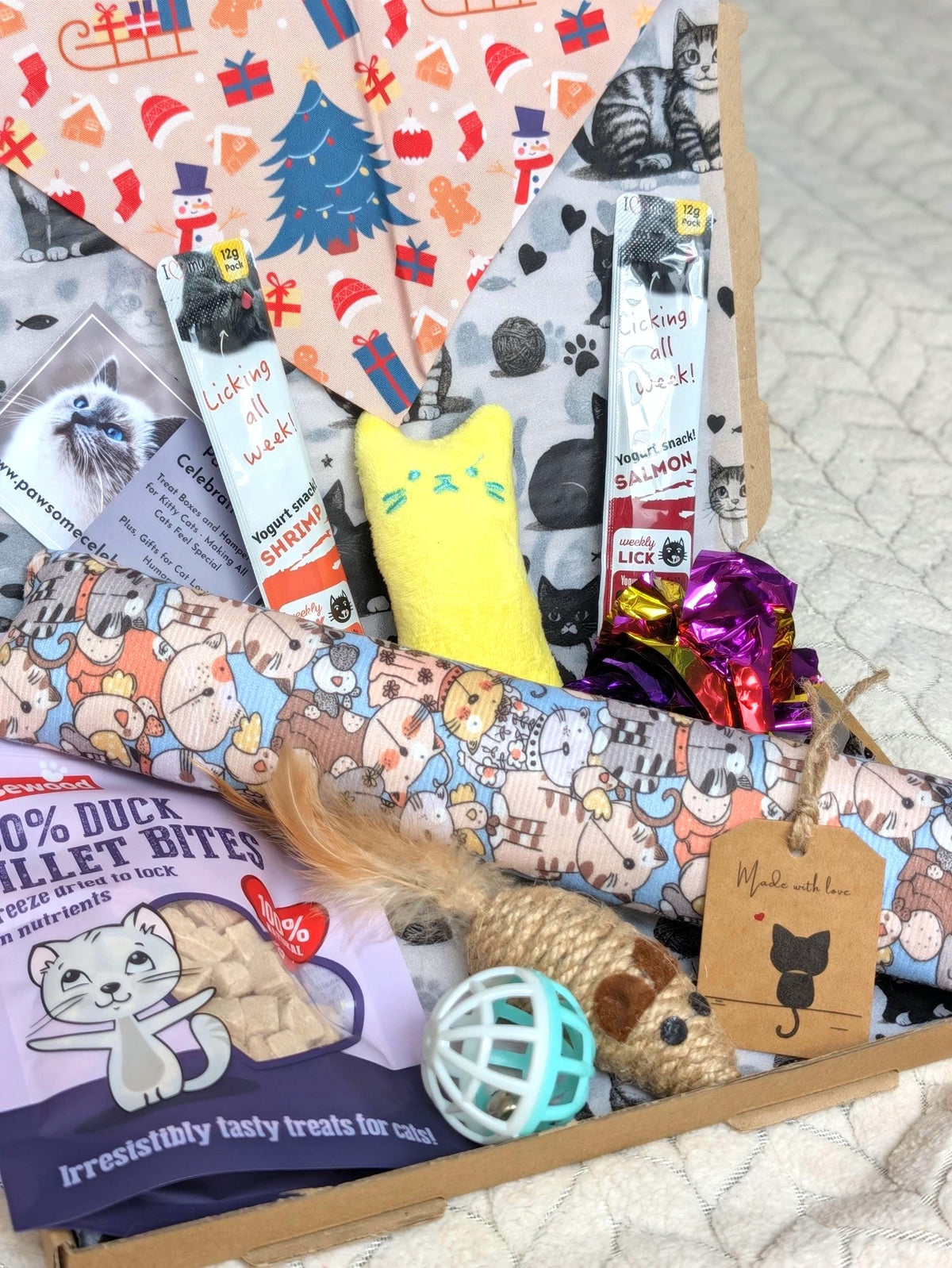 A curated pet gift box filled with various pet treats, toys, and accessories, including a yellow plush toy, a blue ball, a cardboard cat tag, and colorful patterned paper.