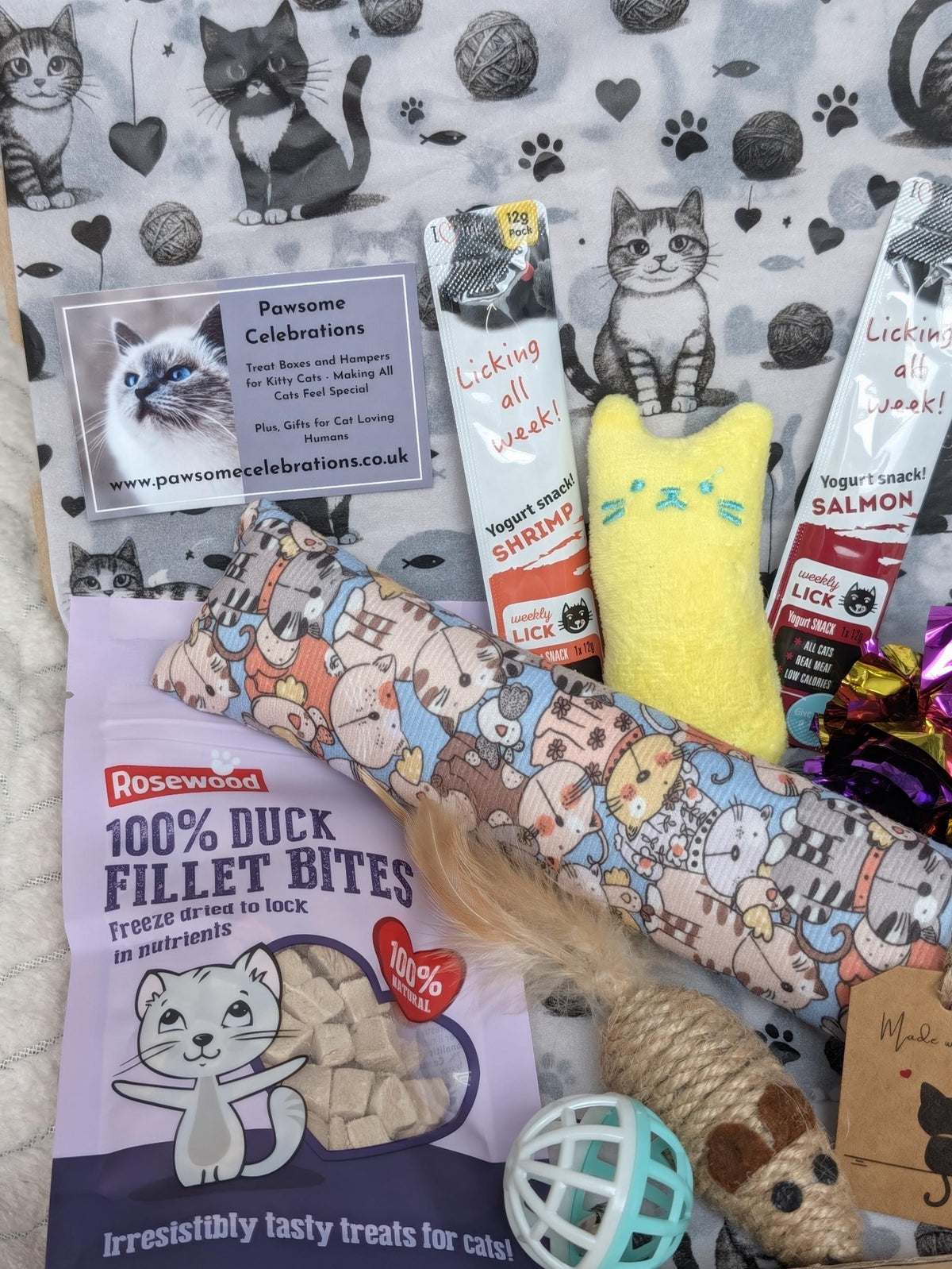 A curated pet gift bundle featuring a pink patterned cat toy, a yellow catnip filled toy, salmon and shrimp flavored treats, a bag of Rosewood duck fillet bites, and a Pawsome Celebrations card, all arranged on a white background with a black and white cat print.