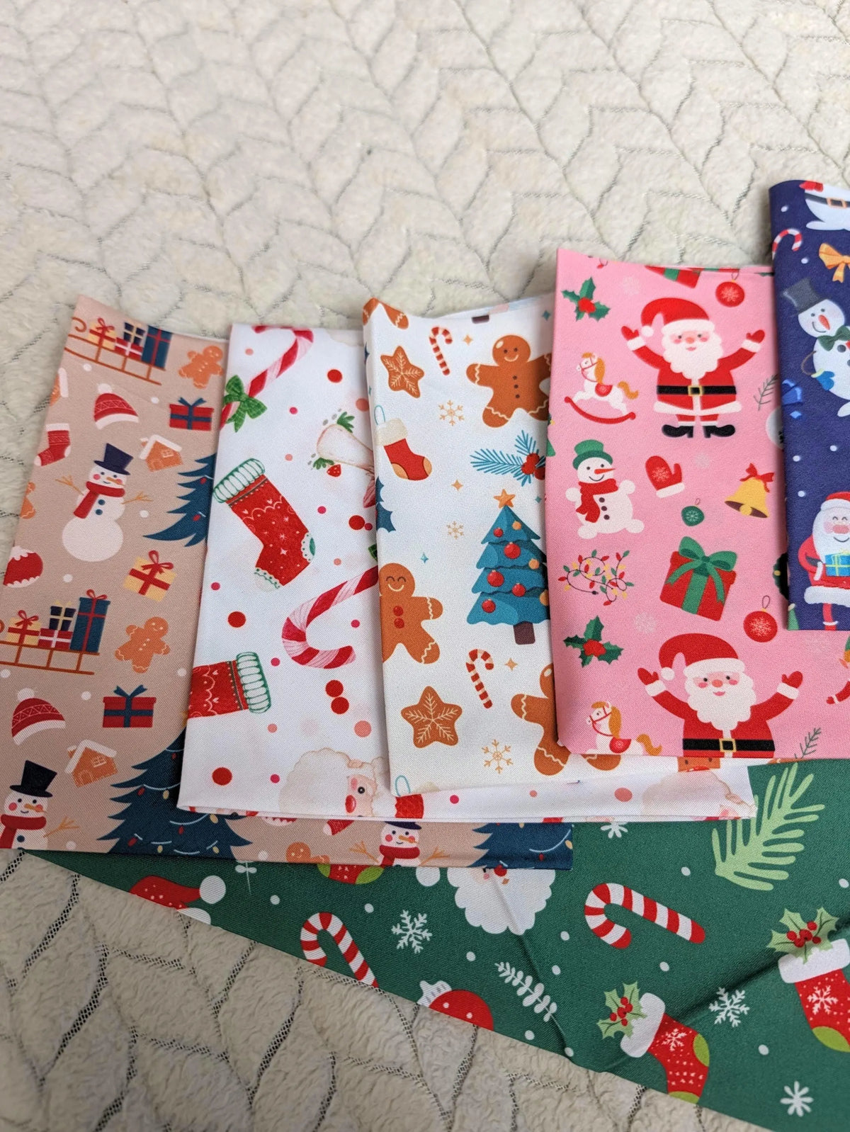 A set of colorful Christmas-themed fabric napkins or small fabric pieces fanned out on a quilted cream surface. The fabrics feature holiday prints including Santa Claus, snowmen, gingerbread men, candy canes, stockings, and trees in pink, white, blue, green, and beige tones.